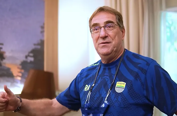 Close-up of Robert Rene Alberts, a middle-aged man with glasses, wearing a blue Persib Bandung jersey and giving a thumbs-up gesture. He appears to be indoors with a blurred background.